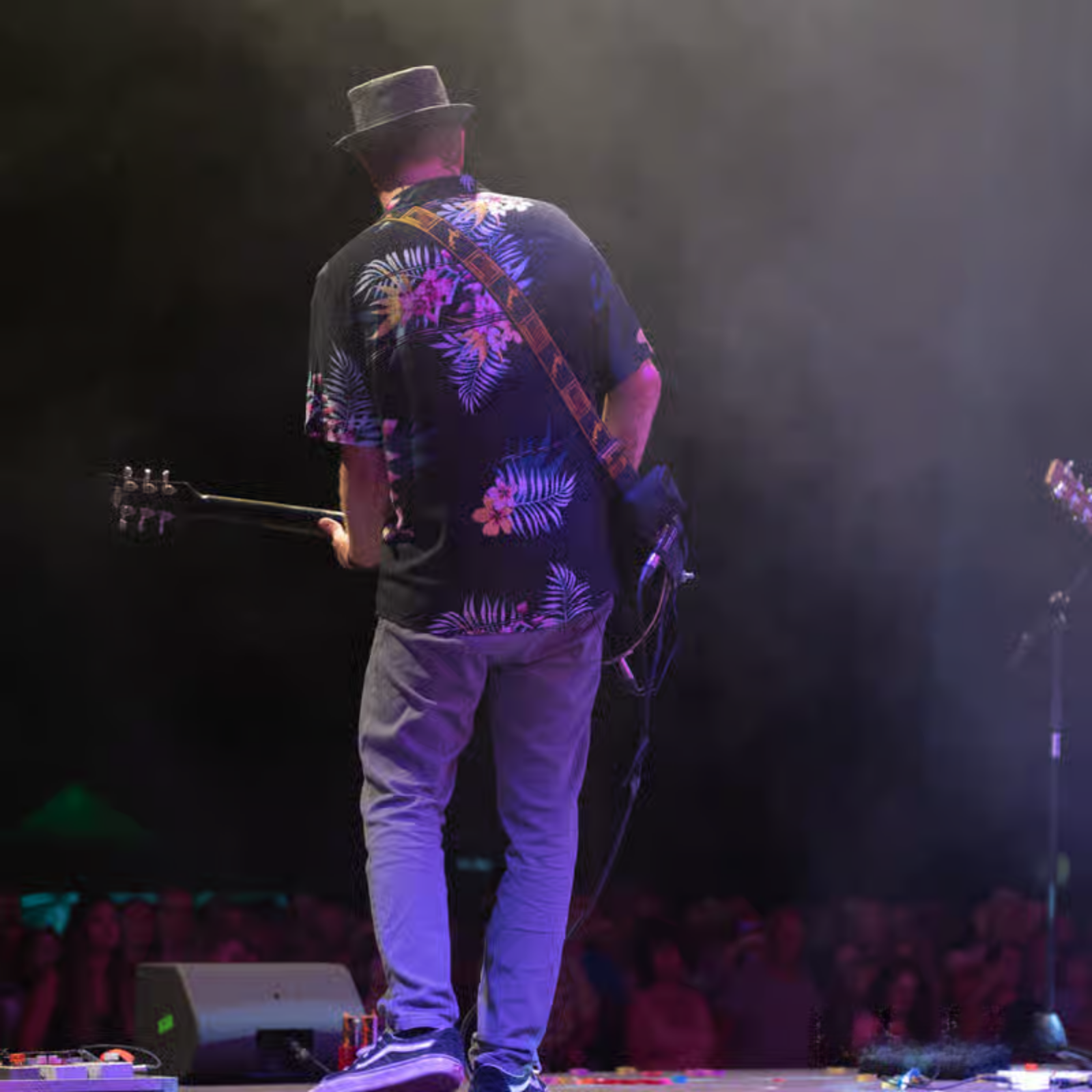 Ein Mann mit einem schwarzen Hut steht auf einer Bühne vor vielen Menschen und spielt Gitarre.