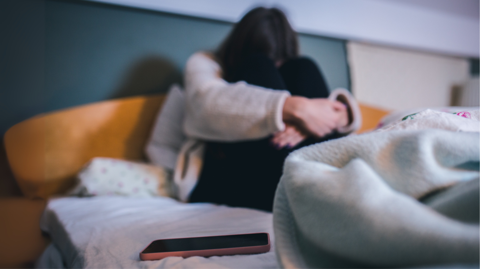 Eine Frau sitzt mit gesenktem Kopf und verschränkten armen aufgelöst im Bett. Im Vordergrund liegt ihr Handy.